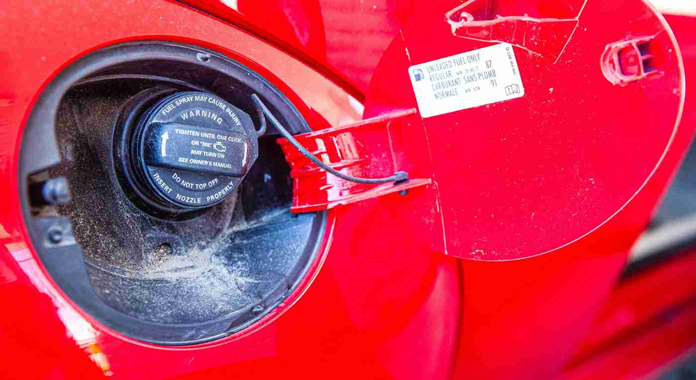 A close-up of a red car’s open fuel door, showing the black fuel cap with warning text and an unleaded fuel label on the inside of the door.