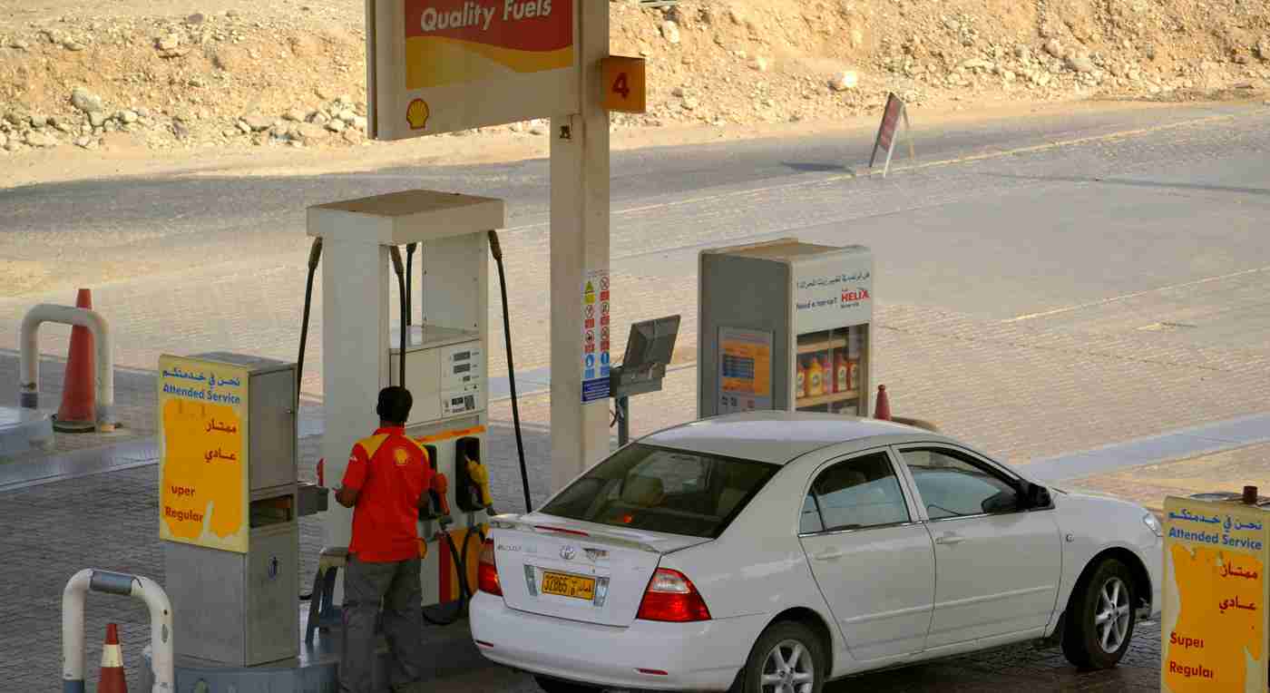 A white car is parked at a fuel pump in a desert gas station. An attendant in a red uniform stands by the pump. Signs display fuel options in English and Arabic.