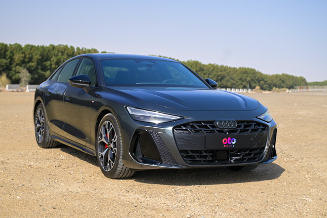 Front angle perspective of a metallic dark grey Audi A6 luxury sedan parked on a sunny gravel terrain.