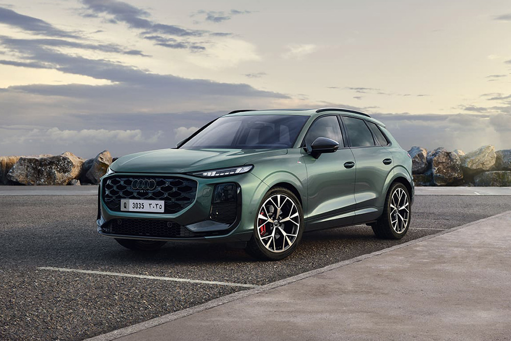 Sleek green Audi Q3 SUV parked on a coastal road, with rocky shoreline and cloudy sky in the background. Modern design with sporty alloy wheels.