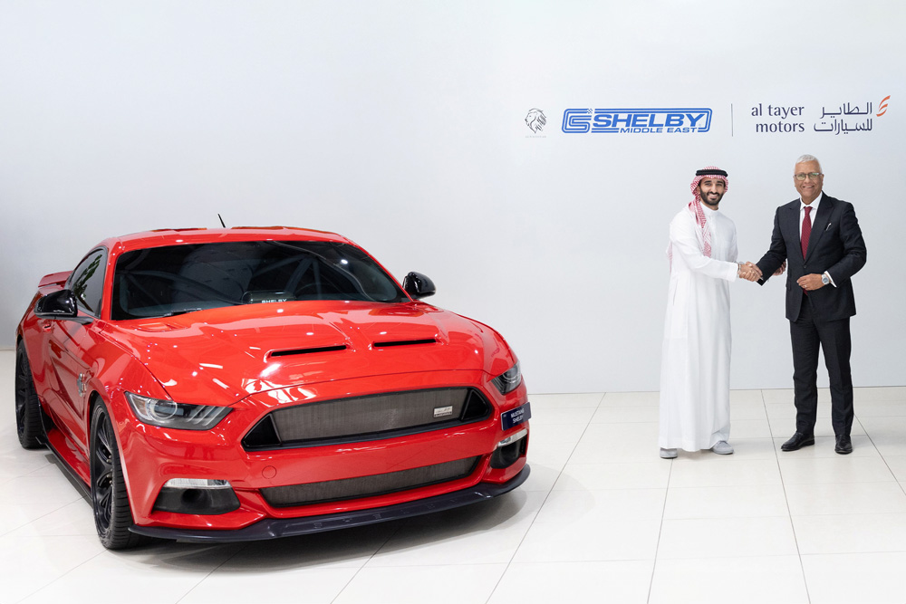Two men in formal attire shake hands next to a red Shelby sports car, Logos for Shelby Middle East and Al Tayer Motors are visible on the wall, conveying a sense of partnership and automotive luxury.