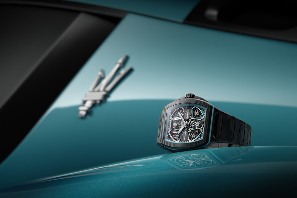 Sleek black watch with an intricate dial sits on a glossy teal car hood. Maserati MCPura performance with Swiss haute horlogerie craftsmanship. A blurred emblem in the background suggests luxury and sophistication.