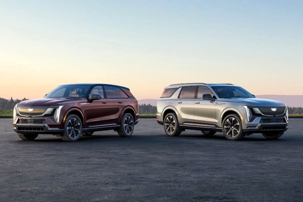Two modern SUVs Escalade IQ & Escalade IQL parked on a scenic open road at sunset. The left SUV is dark red, the right is silver. Both feature sleek designs with large wheels.
