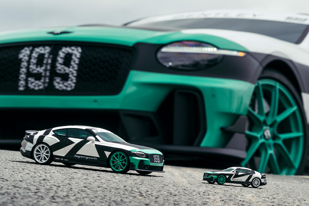 Two toy race cars with black and white patterns and green accents are on a textured surface. A large car with matching design is in the blurred background.