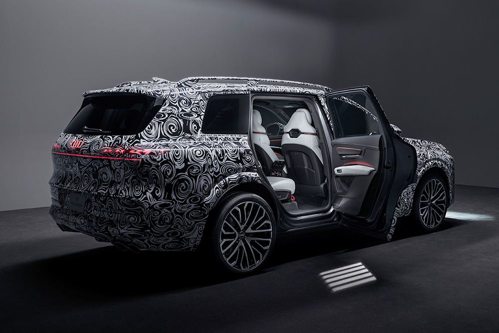 Camouflaged Audi Q9 SUV in a dimly lit room with an open door revealing a white and gray interior. The exterior features a swirl pattern, evoking curiosity.