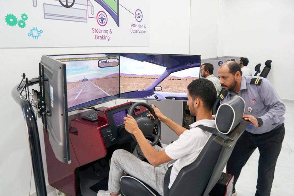 A man in a driving simulator sits at the wheel, focused on a screen showing a road. An instructor observes closely, with tools visible in the room.