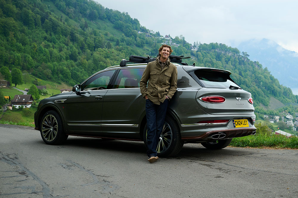 Man smiling, leaning on a green Bentley Bentayga SUV parked on a scenic winding road. Lush green hills and cloudy sky in the background convey a serene vibe.