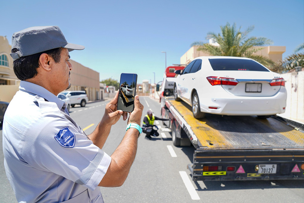 A uniformed man takes a photo of a white car on a tow truck under a clear blue sky. The scene depicts roadside assistance on a suburban street.