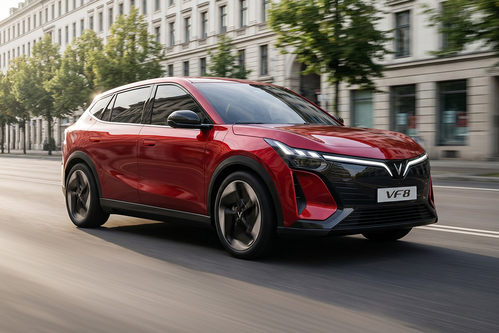 A metallic crimson red VinFast VF 8 electric crossover SUV driving smoothly along a European city street during the day.