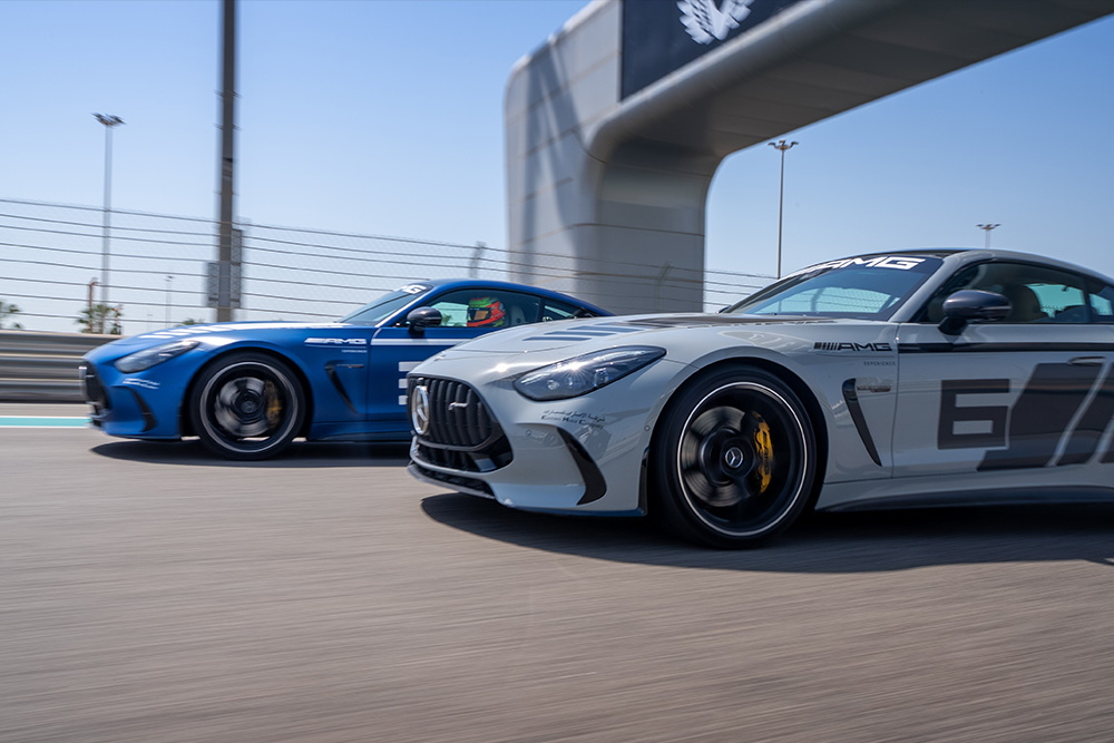 Two high-performance sports cars—one silver and one blue—race side by side under clear skies, capturing intense speed and competition as Yas Marina Circuit launches the Mercedes-AMG experience.