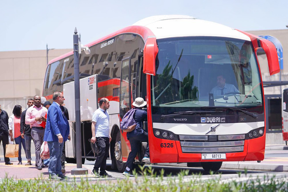 A group of people boards a modern red and white city bus on a sunny day. The bus, marked "Dubai," is parked at a bus stop, suggesting a public transportation setting. People appear relaxed and organized.