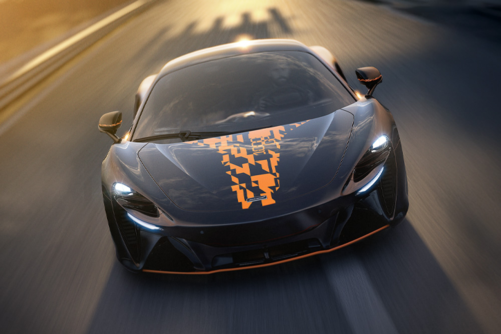 A sleek black McLaren Artura sports car with digital orange decals on the hood speeds down a blurred racetrack, headlights on, conveying a sense of speed and precision.