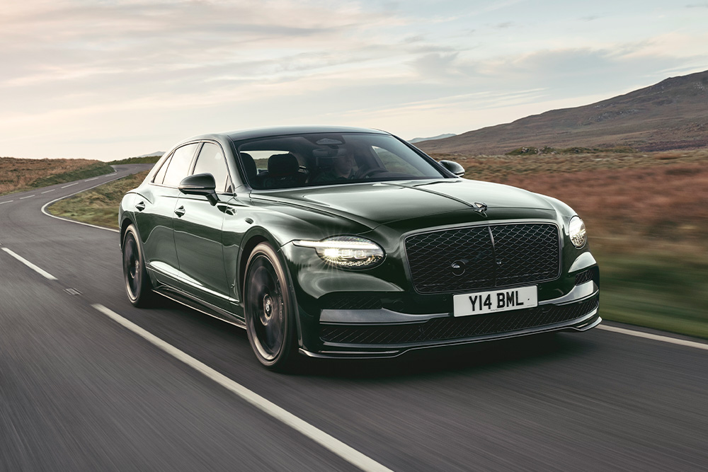 A sleek, dark green luxury Bentley Spur car driving on a winding rural road with hills in the background, conveying sophistication and power on a serene journey.