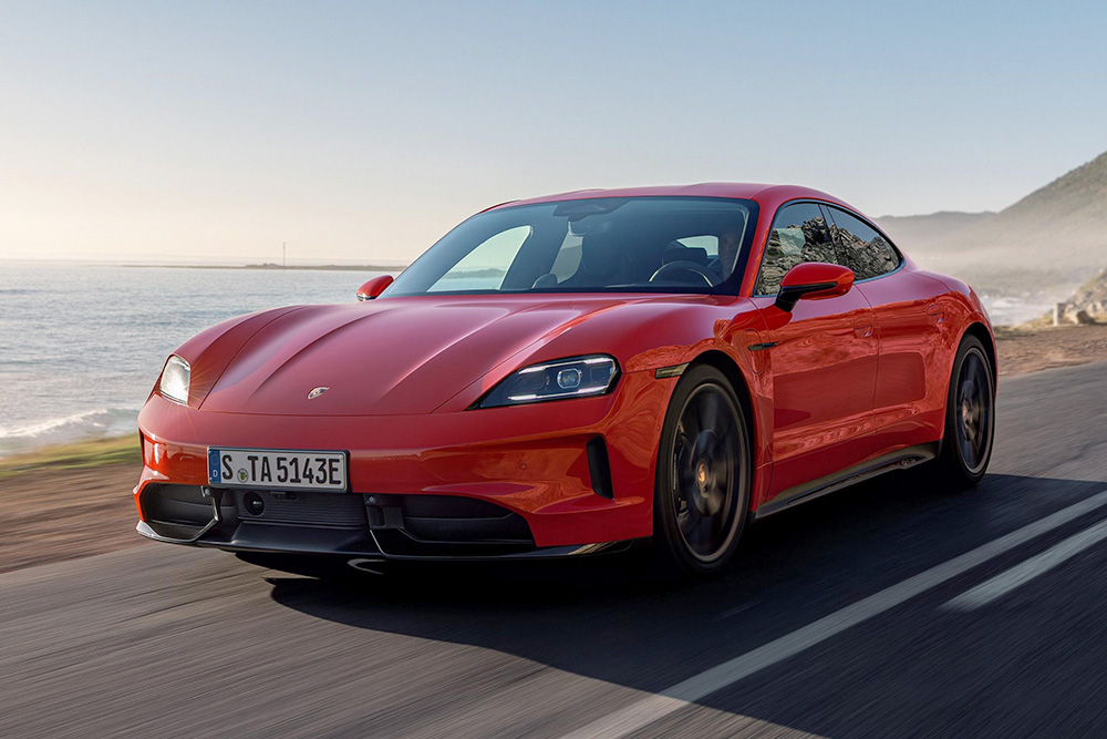 Front three-quarter view of a bright red Porsche Taycan electric sedan cruising along a coastal road.
