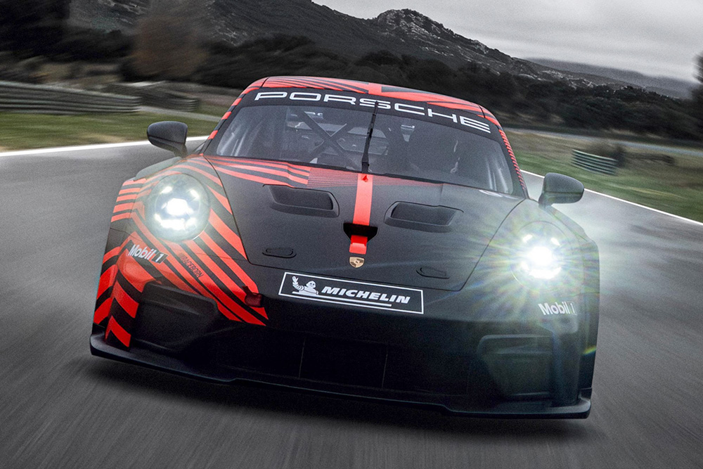 Black Porsche 911 GT3 race car with red stripes driving fast on a race track with headlights shining.