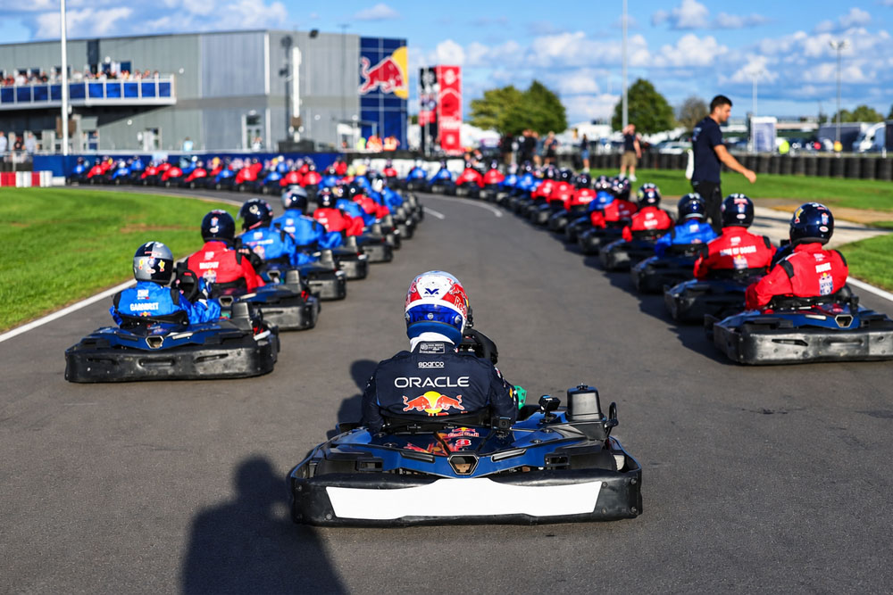 Go-kart drivers lined up in branded racing suits on an outdoor asphalt circuit